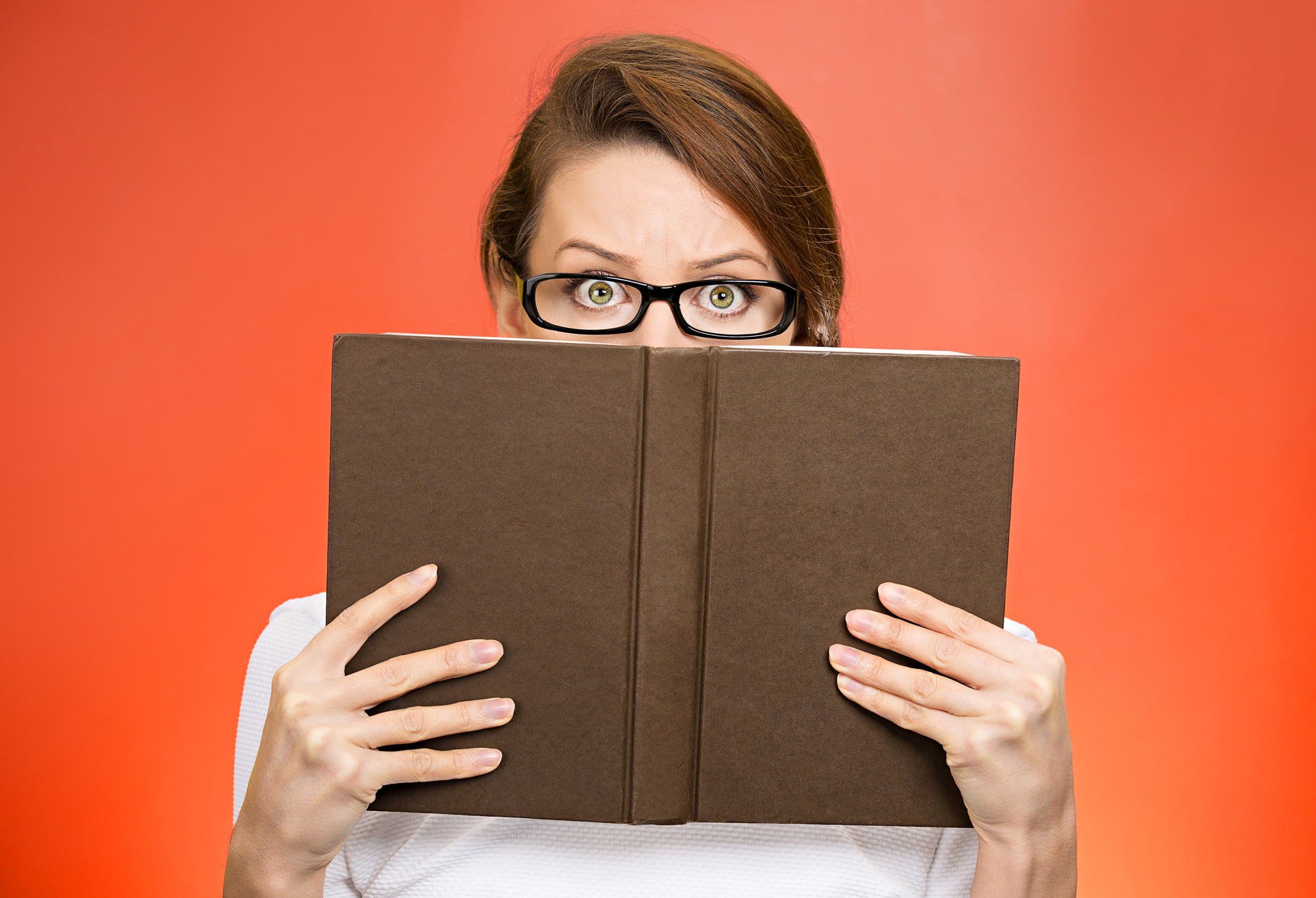 Closeup portrait of woman with glasses looking over a book with a red background.